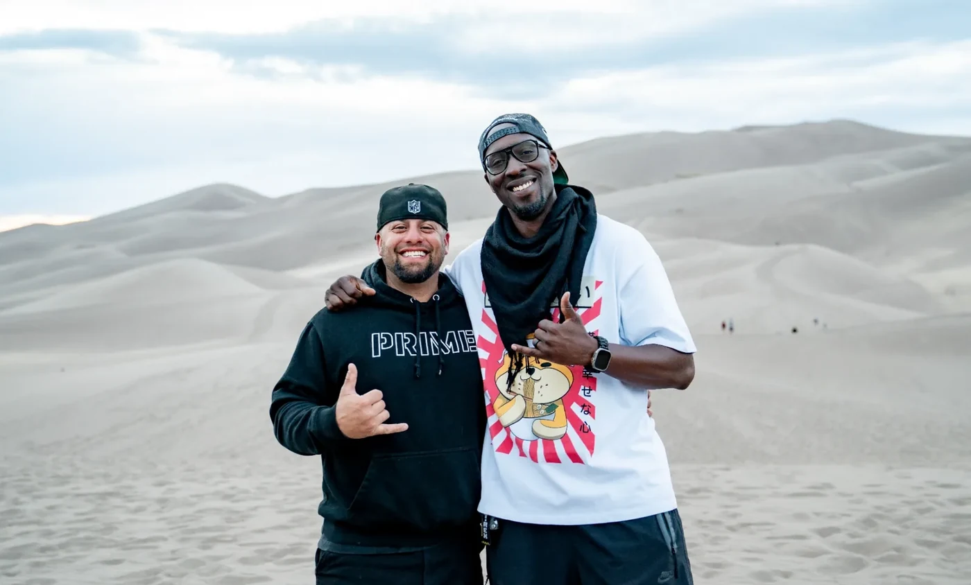 Ian Bennett and Cory Corrizalez on the Great Sand Dunes at golden hour, arms around each other, both throwing shakas — twenty years of friendship, one shoot day